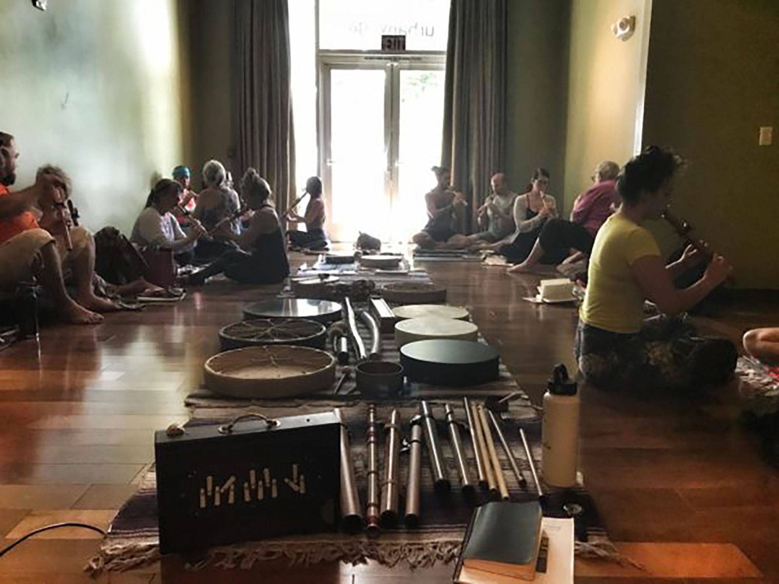A training group sitting in a circle on a studio floor, each person holding a flute, with a shruti box, frame drums, chimes and mallets laid out on rugs between them