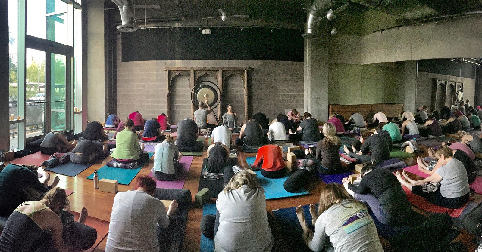 A long studio room filled with people lying on mats and blankets during a sound meditation, gongs on stands at the far wall
