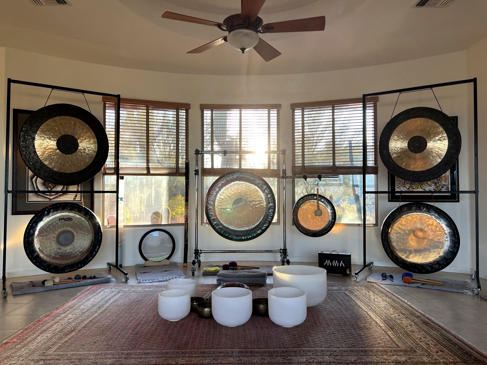 Six gongs hanging on stands around the curved window wall of the room, crystal and Himalayan bowls on a rug in the middle of the floor, morning sun coming through the blinds