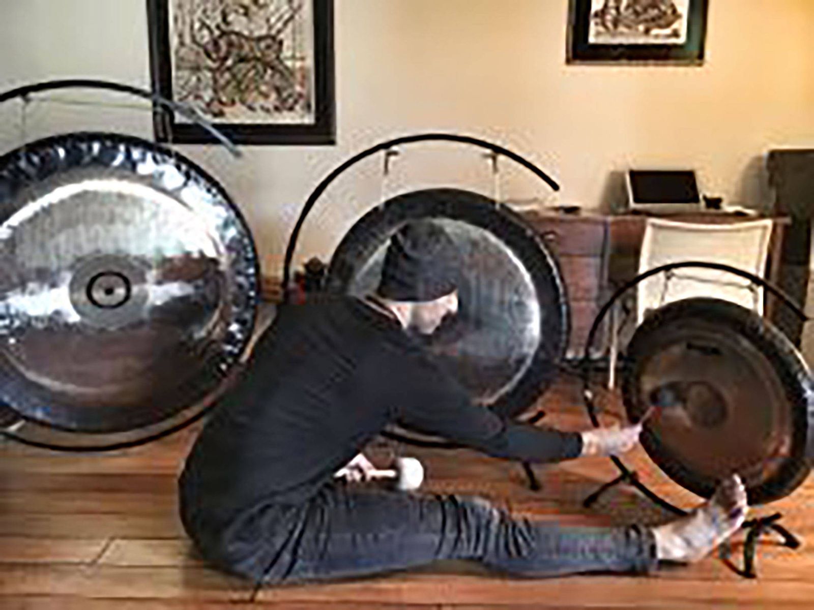 Hamid sitting on a wooden floor among three gongs on stands, reaching out to play the smallest of them with a mallet