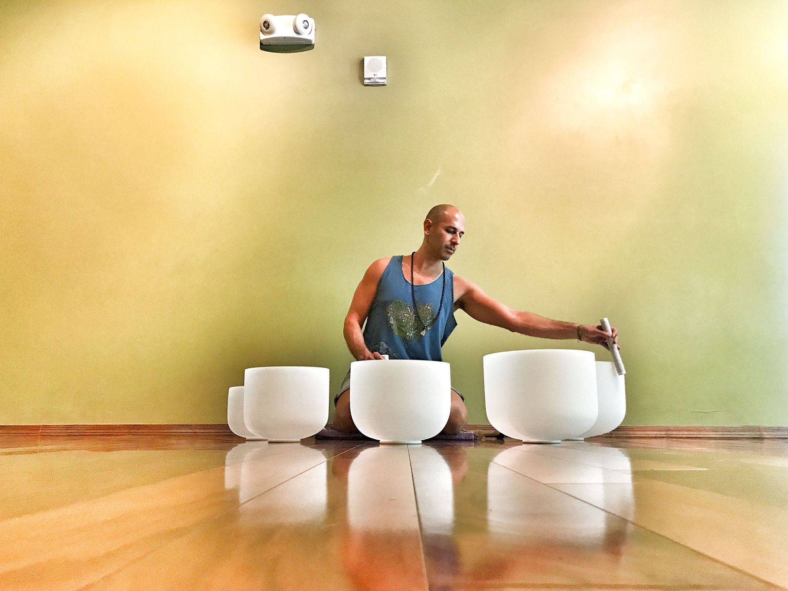 Hamid kneeling on a polished wooden floor among a row of frosted crystal singing bowls, circling the rim of one with a wand, a pale green wall beyond him