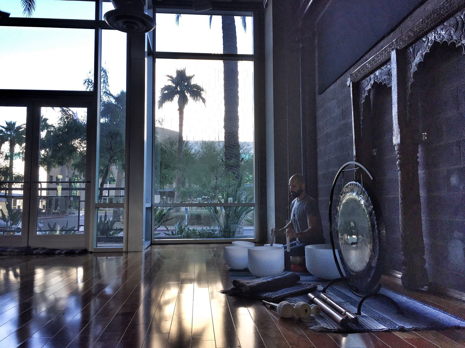 Hamid sitting on the floor of a window lit studio playing crystal singing bowls, a large gong on a stand beside him and palm trees outside the tall windows