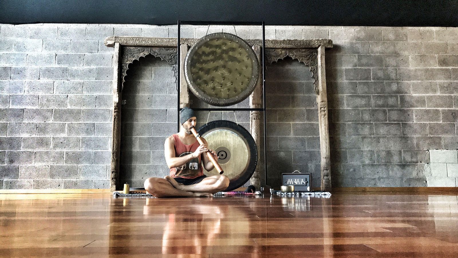 Hamid sitting cross legged on a studio floor playing a flute, two gongs on a carved wooden stand behind him