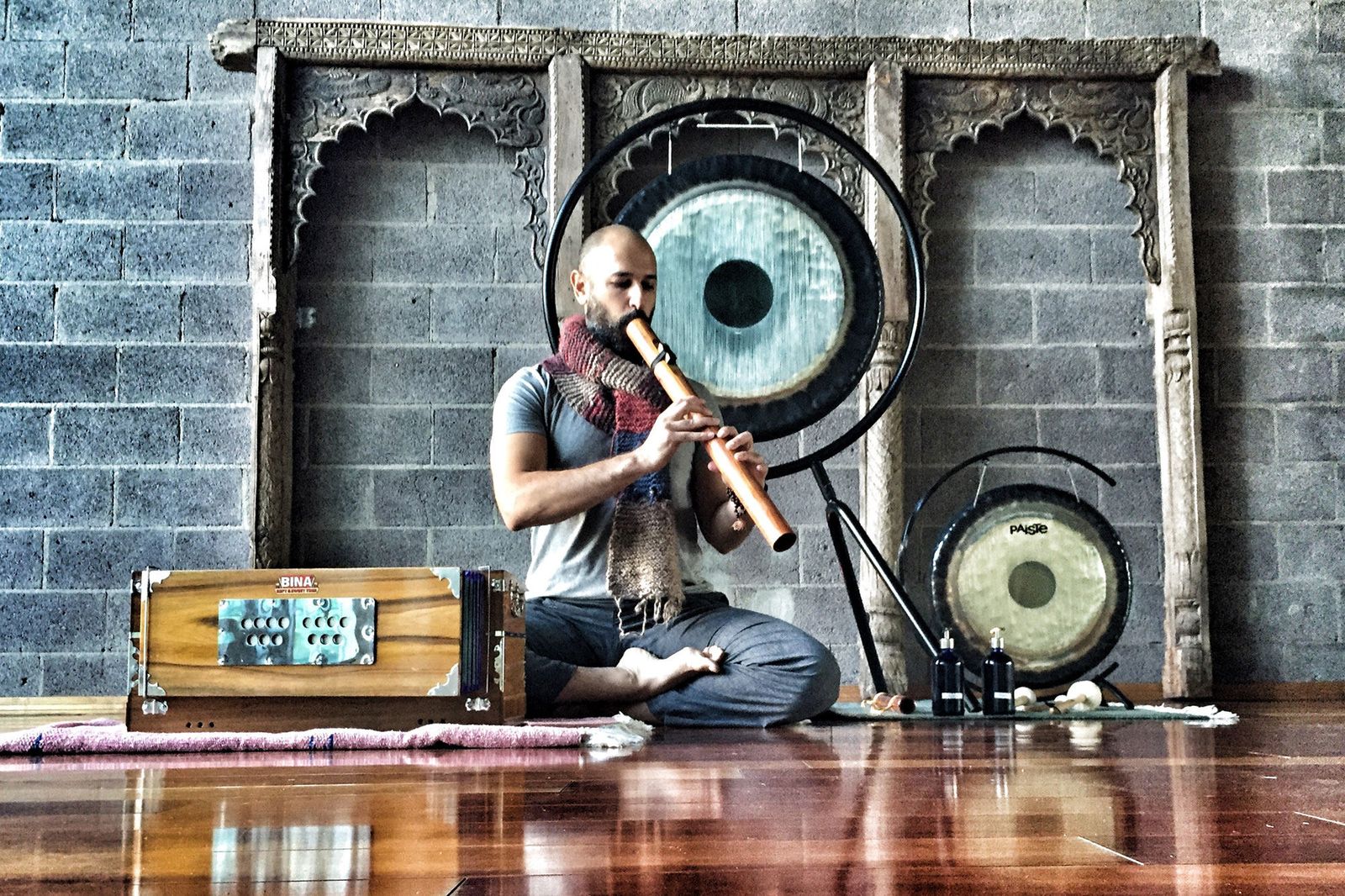 Hamid sitting cross legged on a studio floor playing a long bamboo flute, a hanging gong behind him, a harmonium on the rug beside him and a second gong to his right