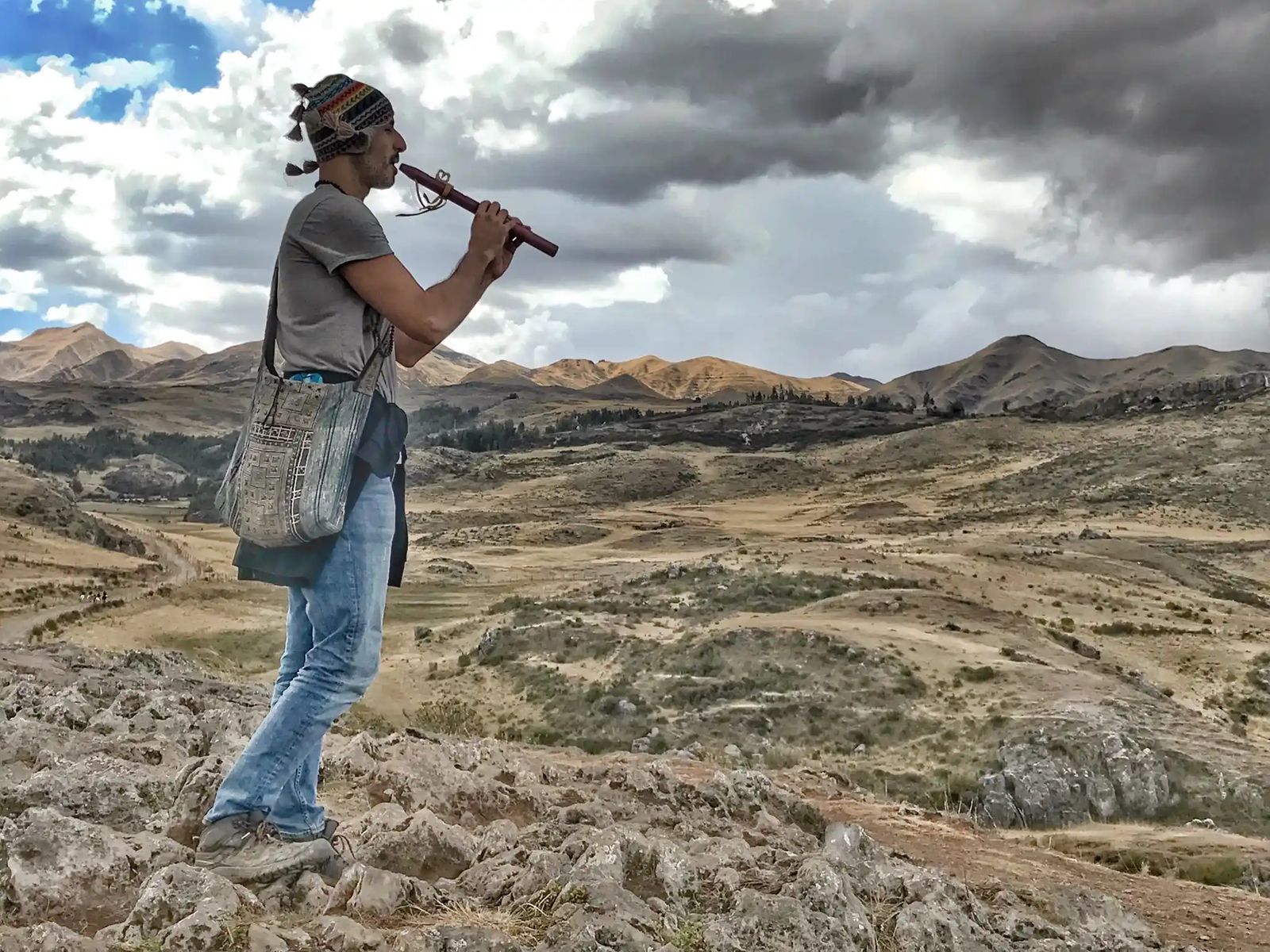 Hamid standing on a rocky hillside playing a wooden flute, a woven bag over his shoulder, dry hills and storm clouds stretching away behind him