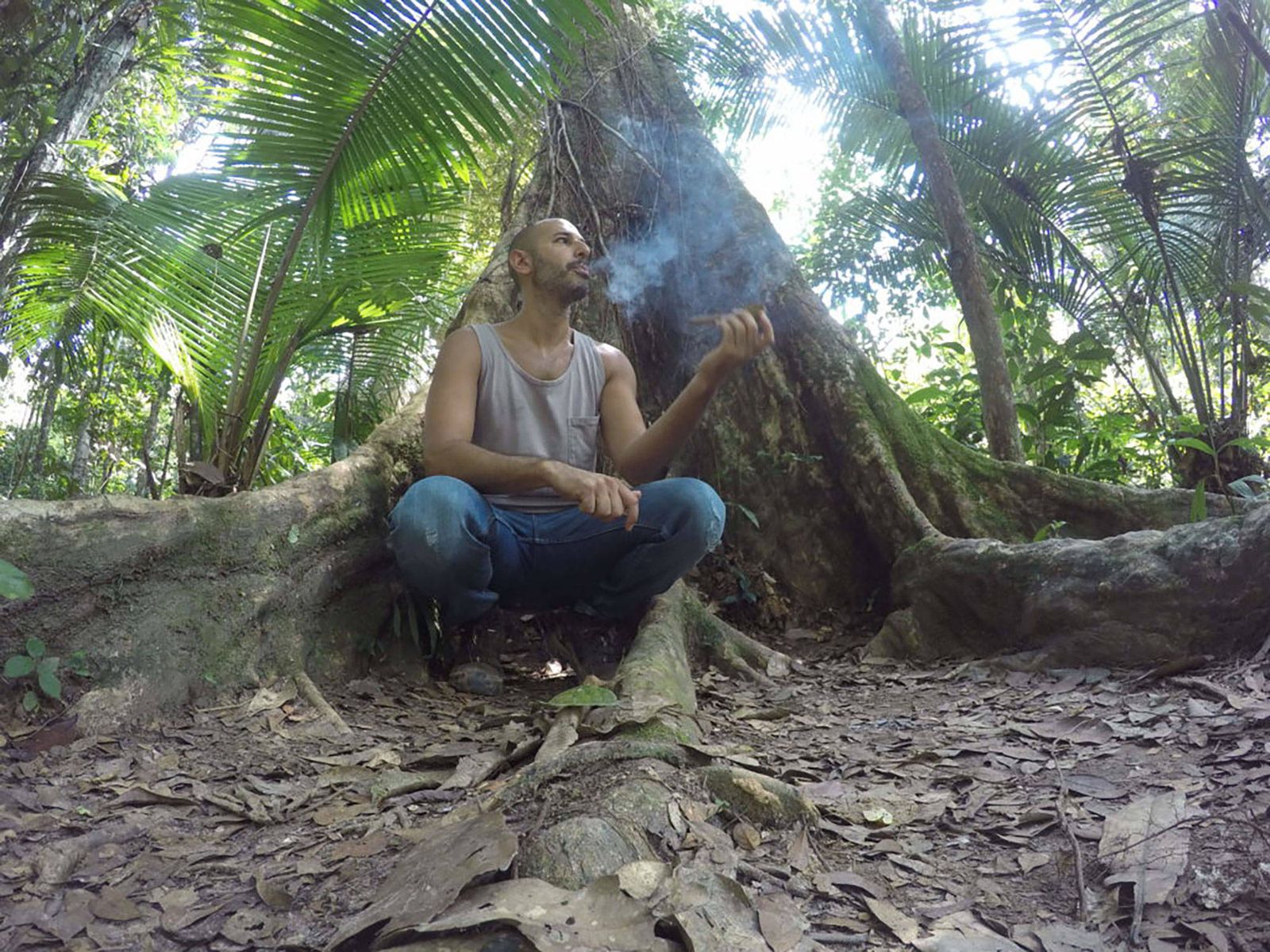 Hamid sitting cross legged in the buttress roots of a large jungle tree in Peru, smoking a mapacho, with smoke rising past his face