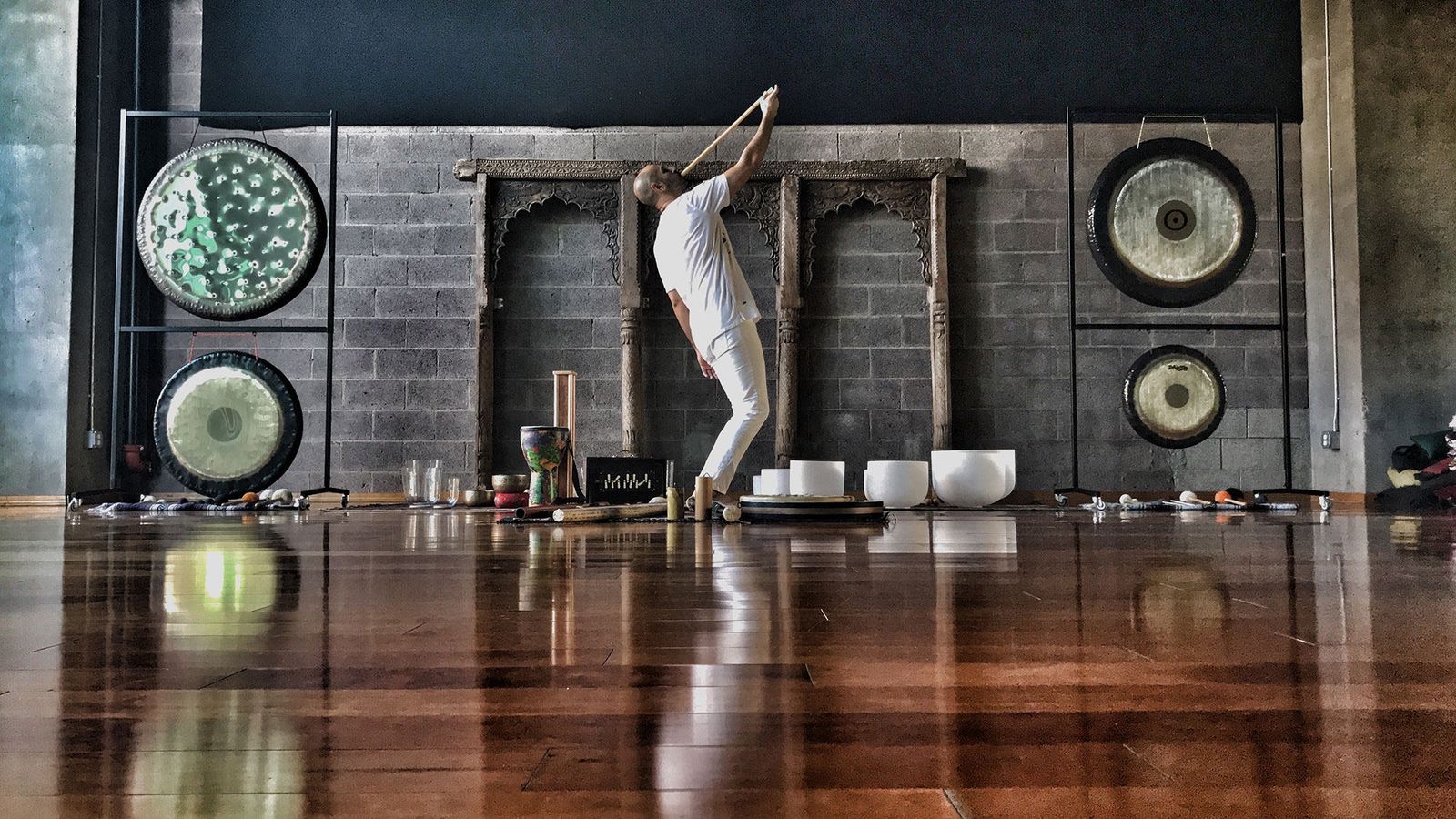 Hamid standing in a studio playing a long overtone flute, with four gongs on stands and a row of crystal bowls along the wall behind him