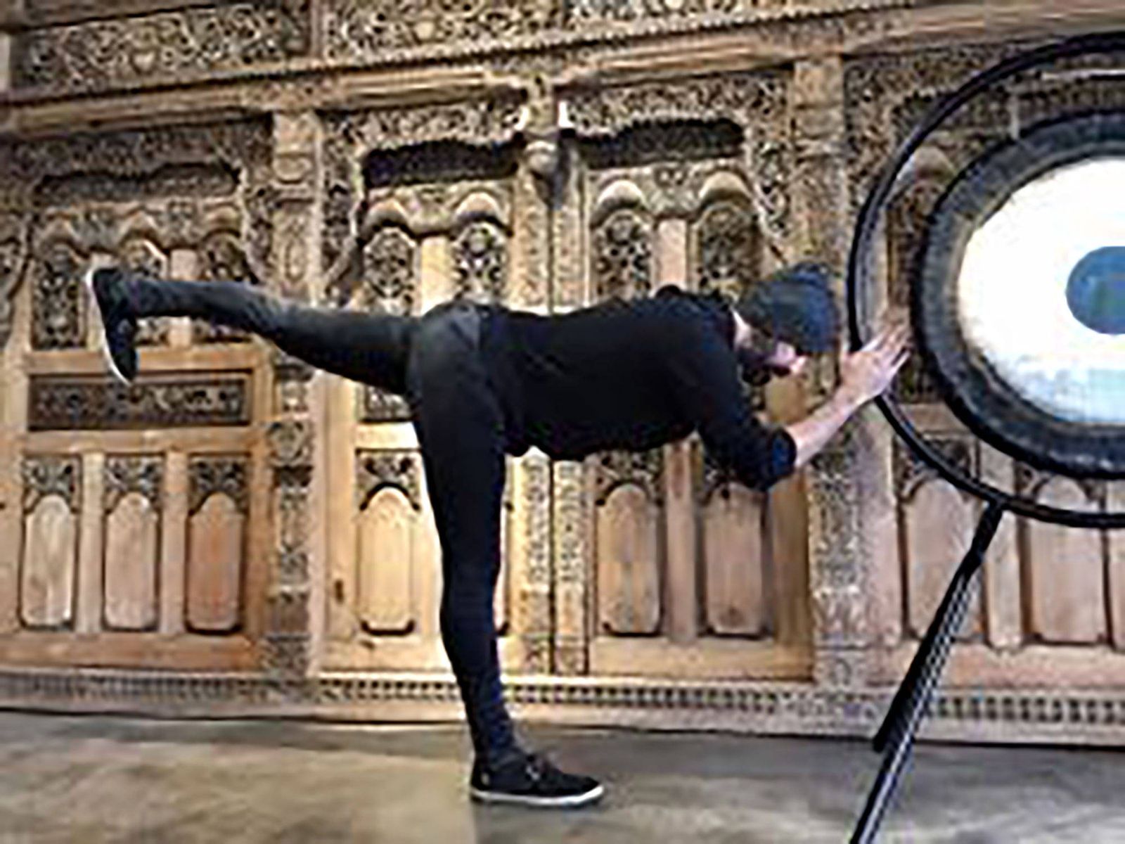 Hamid balancing on one leg with the other stretched out behind him, one hand on the rim of a large gong on its stand, in front of a carved wooden wall