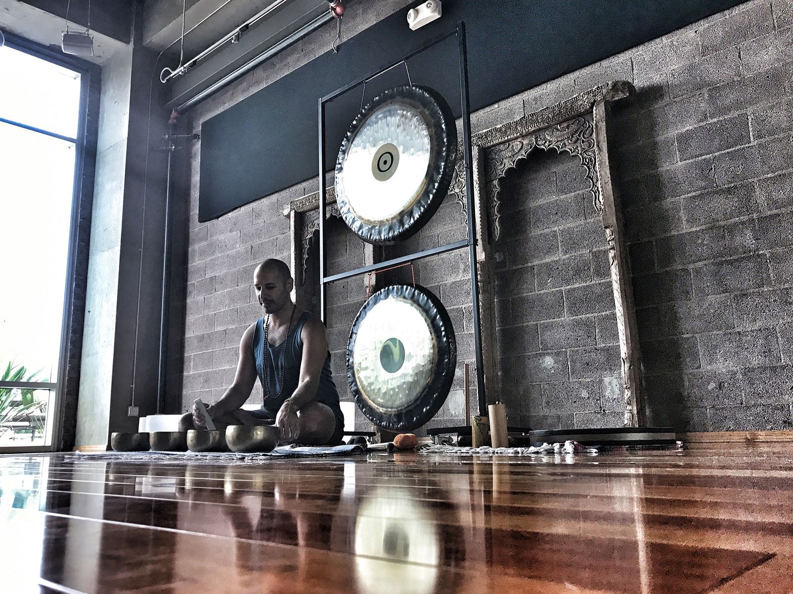 Hamid seated on a studio floor with a row of Himalayan bowls in front of him and two large gongs hanging on a stand beside him