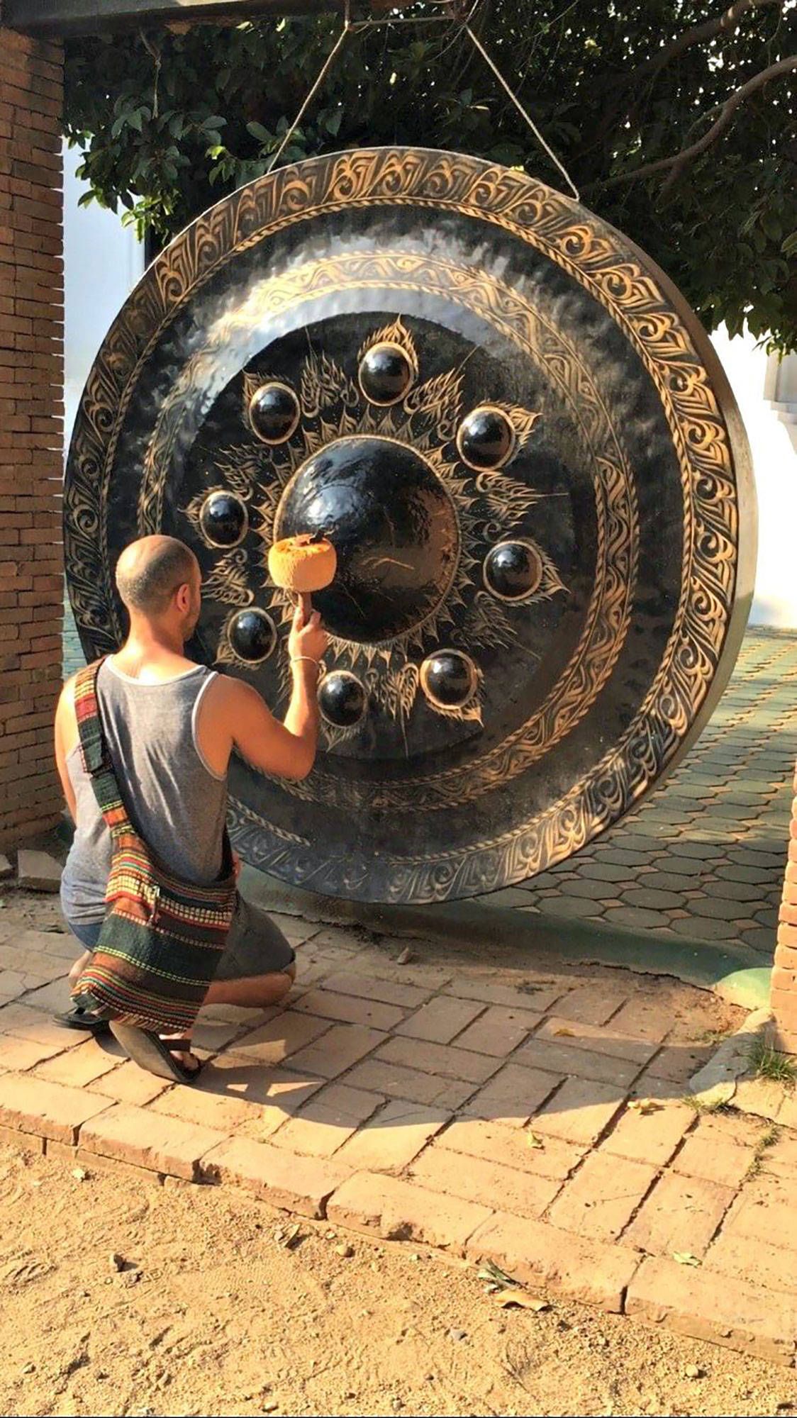 Hamid kneeling to play a large ornate Thai gong hanging outdoors, its face covered in raised bosses and carved detail