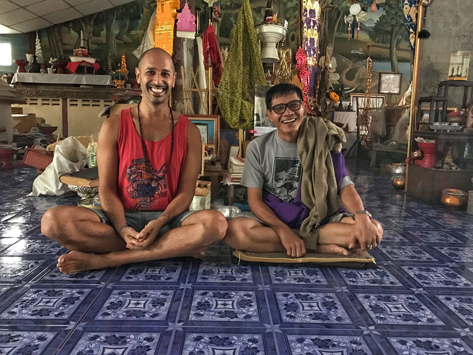 Hamid and Ajahn Pichest Boonthumme sitting cross legged side by side on a patterned tile floor, both smiling, an altar and a crowded wall behind them