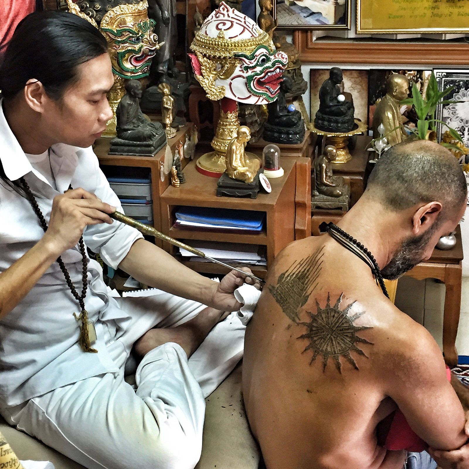 A Thai master tattooing a sak yant on Hamid's back with a traditional rod, in a room full of Buddha figures