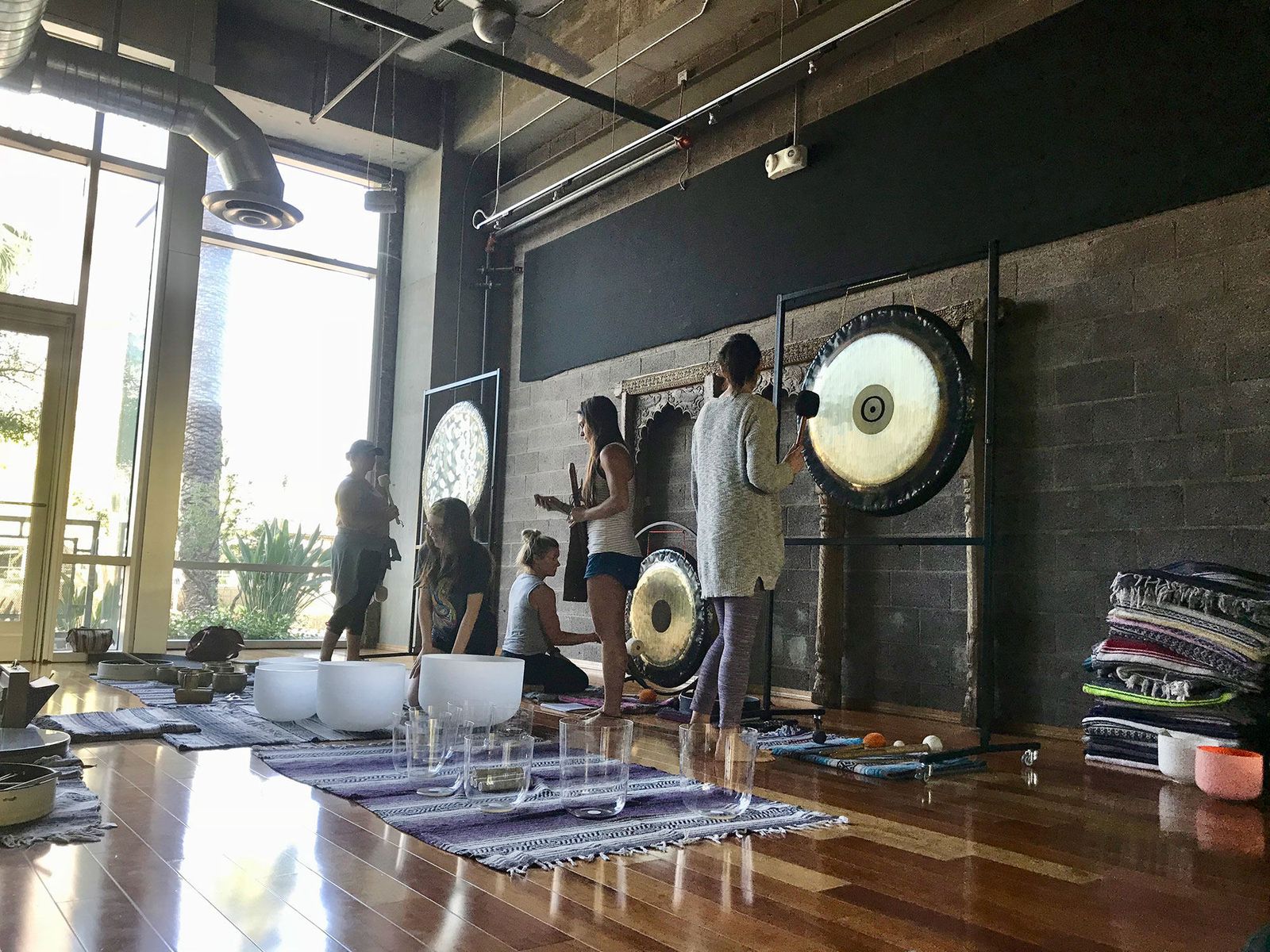 Four students standing at hanging gongs in a studio, playing them with mallets, crystal bowls on the floor in front of them
