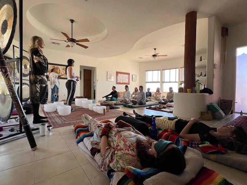 A sound session in progress during a training, people lying down under blankets in the foreground, a row of students seated behind them, crystal bowls on a rug and gongs on stands along the wall