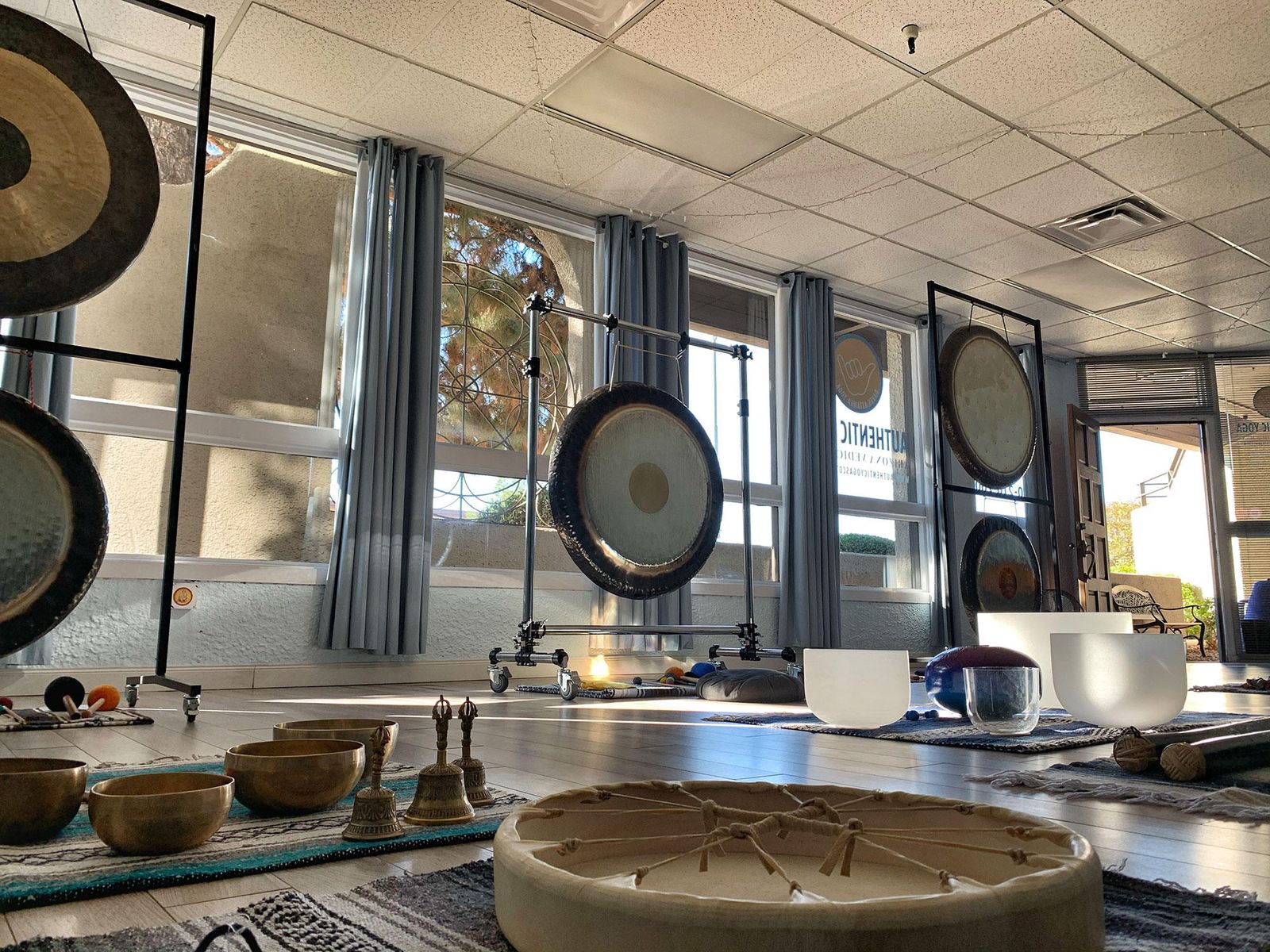 A studio laid out for a training, gongs on stands along a wall of windows, Himalayan bowls, crystal bowls, bells and a frame drum on rugs across the floor